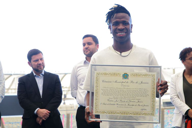 Rio de Janeiro (RJ) -  A sanção da nova lei, de autoria do deputado Professor Josimar (Psol), aconteceu durante evento no Maracanã, que contou com a presença do próprio Vini Jr. Ele foi homenageado com a Medalha Tiradentes, maior prêmio concedido pela Assembleia Legislativa do Estado do Rio de Janeiro (Alerj), com a Medalha Pedro Ernesto e com o título de Cidadão Carioca, concedido pela Câmara Municipal do Rio. O atleta também gravou os pés na “Calçada da Fama” do Maracanã. 
Foto:Governo do Estado/RJ/Divulgação 